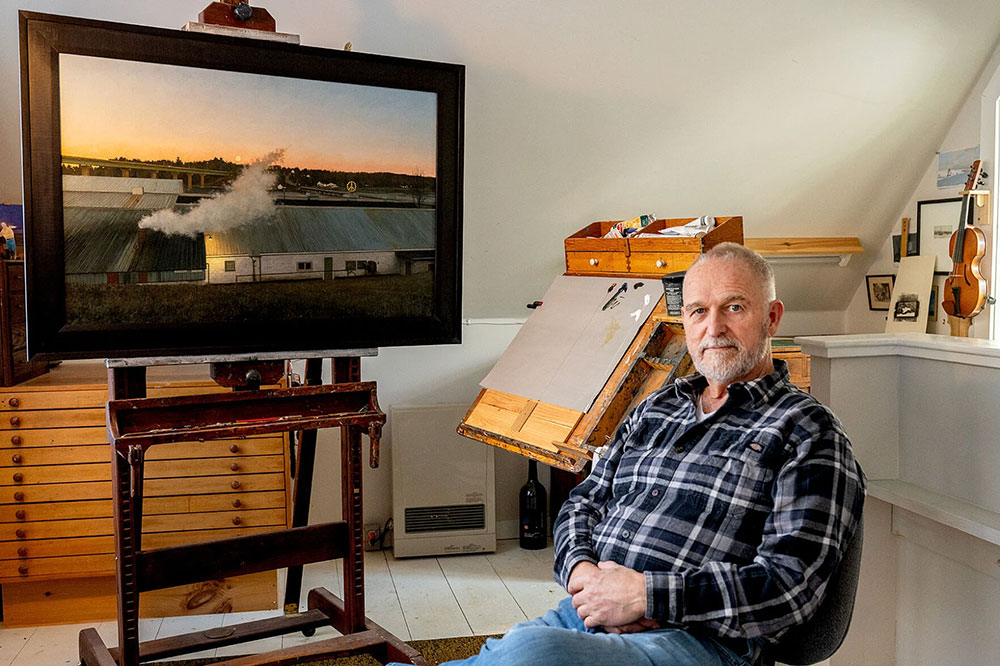 Click to enlarge A man casually sits in a chair next to a realistic painting of a white building with smoke rising above it.